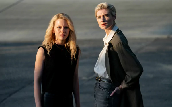 HD desktop wallpaper of movie Maxxxine featuring Mia Goth and Elizabeth Debicki. They stand outdoors, bathed in soft sunlight, projecting intense expressions.