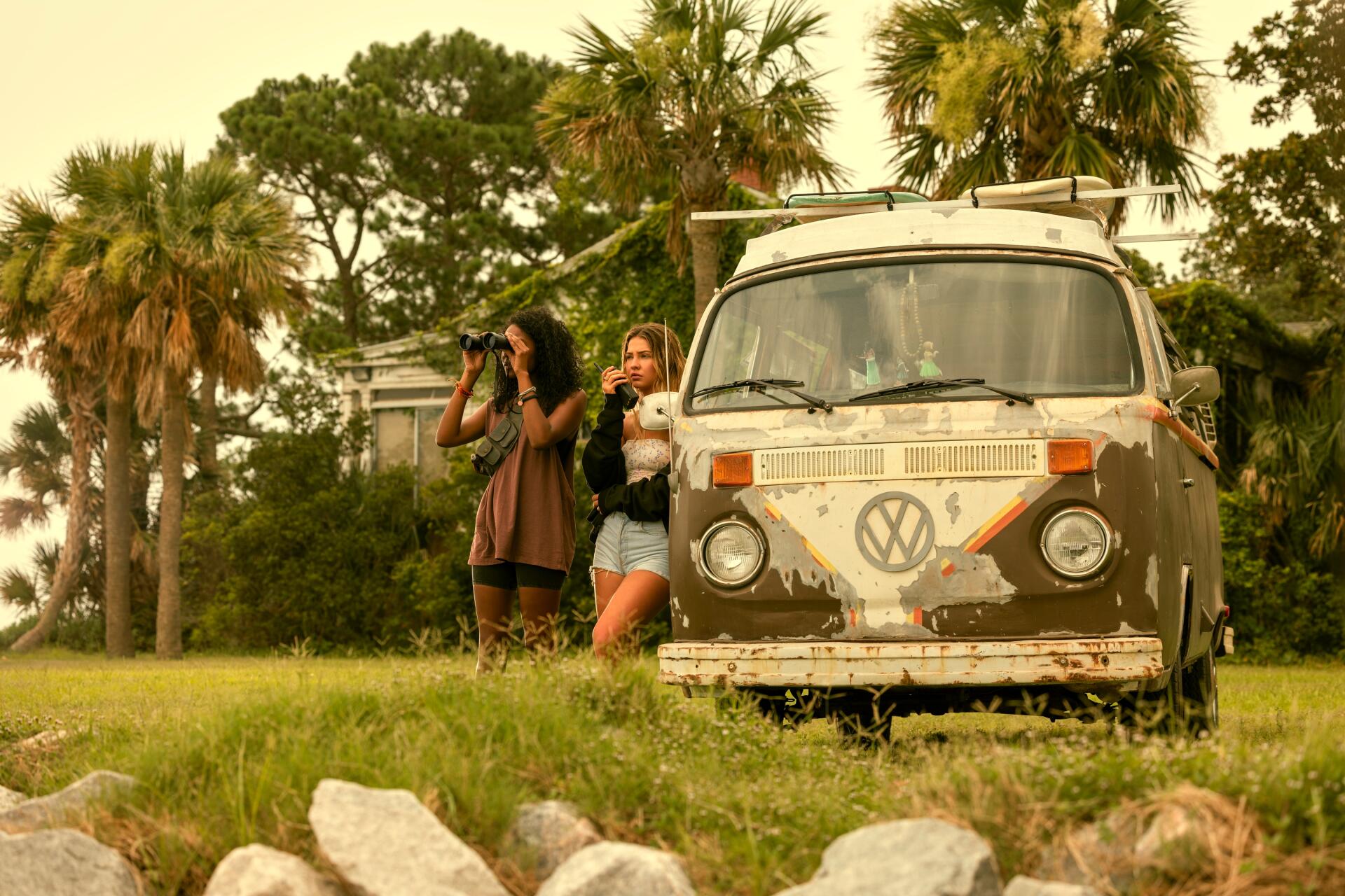 Two characters from the TV show Outer Banks explore a lush, beachside scene, with one using binoculars next to a vintage Volkswagen van. A vibrant, adventurous backdrop captures the essence of the series.