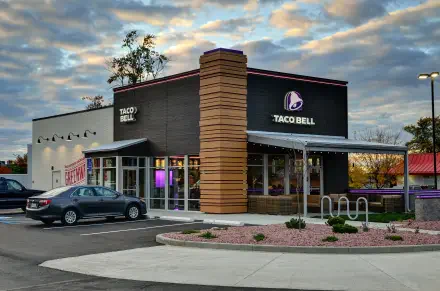 A modern Taco Bell restaurant with a distinctive design, set against a dramatic sky. The image captures the building's architecture and surrounding area in vibrant detail.