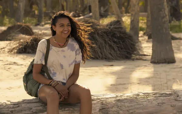 Madison Bailey smiles while sitting on a log in a beach setting from the TV show Outer Banks. The image captures the show's adventurous spirit in vibrant HD resolution.