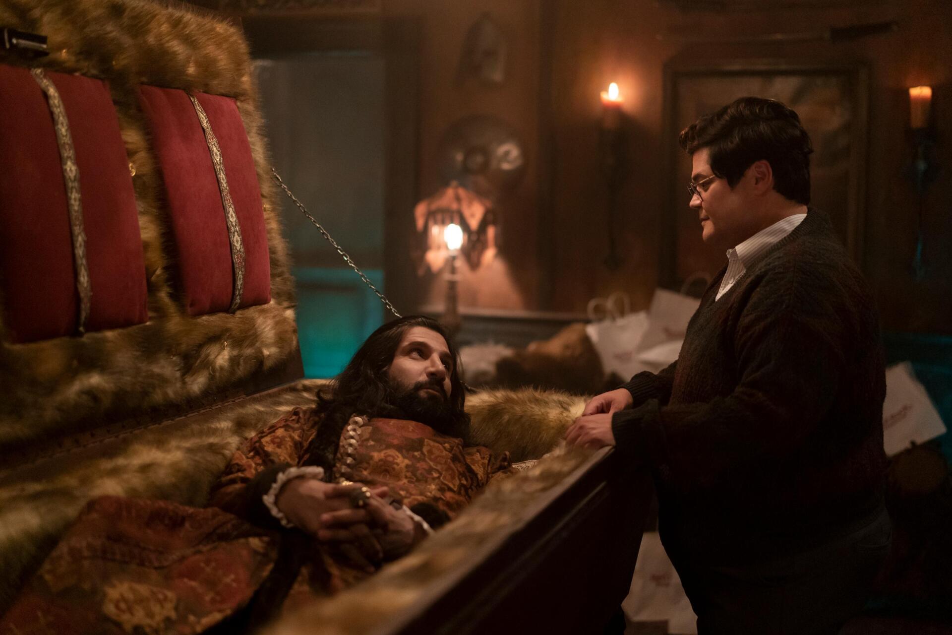 A scene from the TV show What We Do in the Shadows, featuring a humorous interaction between a vampire in a coffin and a human character, set in a dimly lit room.