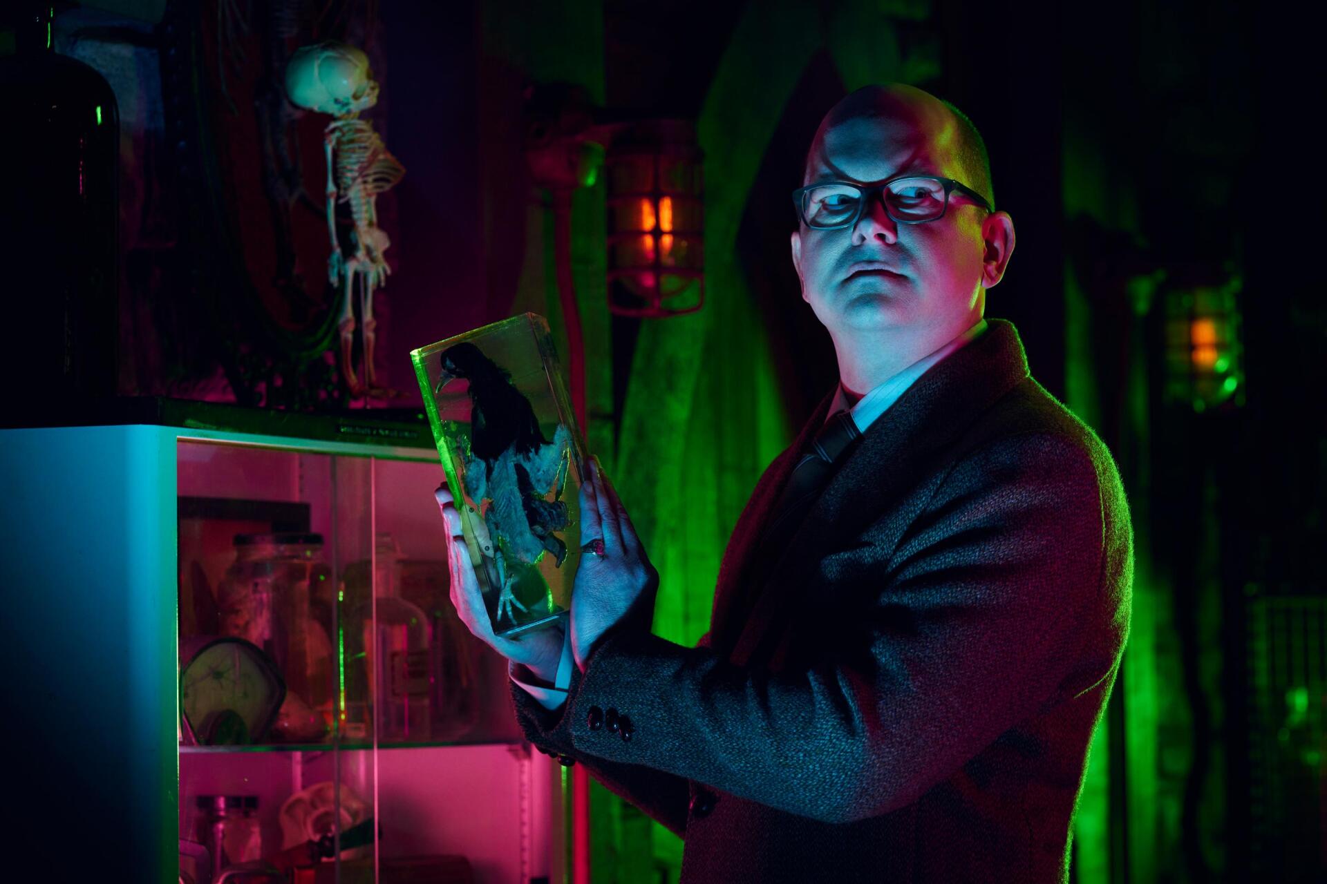 Mark Proksch stands dramatically in a colorful, eerie setting from the TV show *What We Do in the Shadows*, holding an object amidst shadows and vibrant lighting.