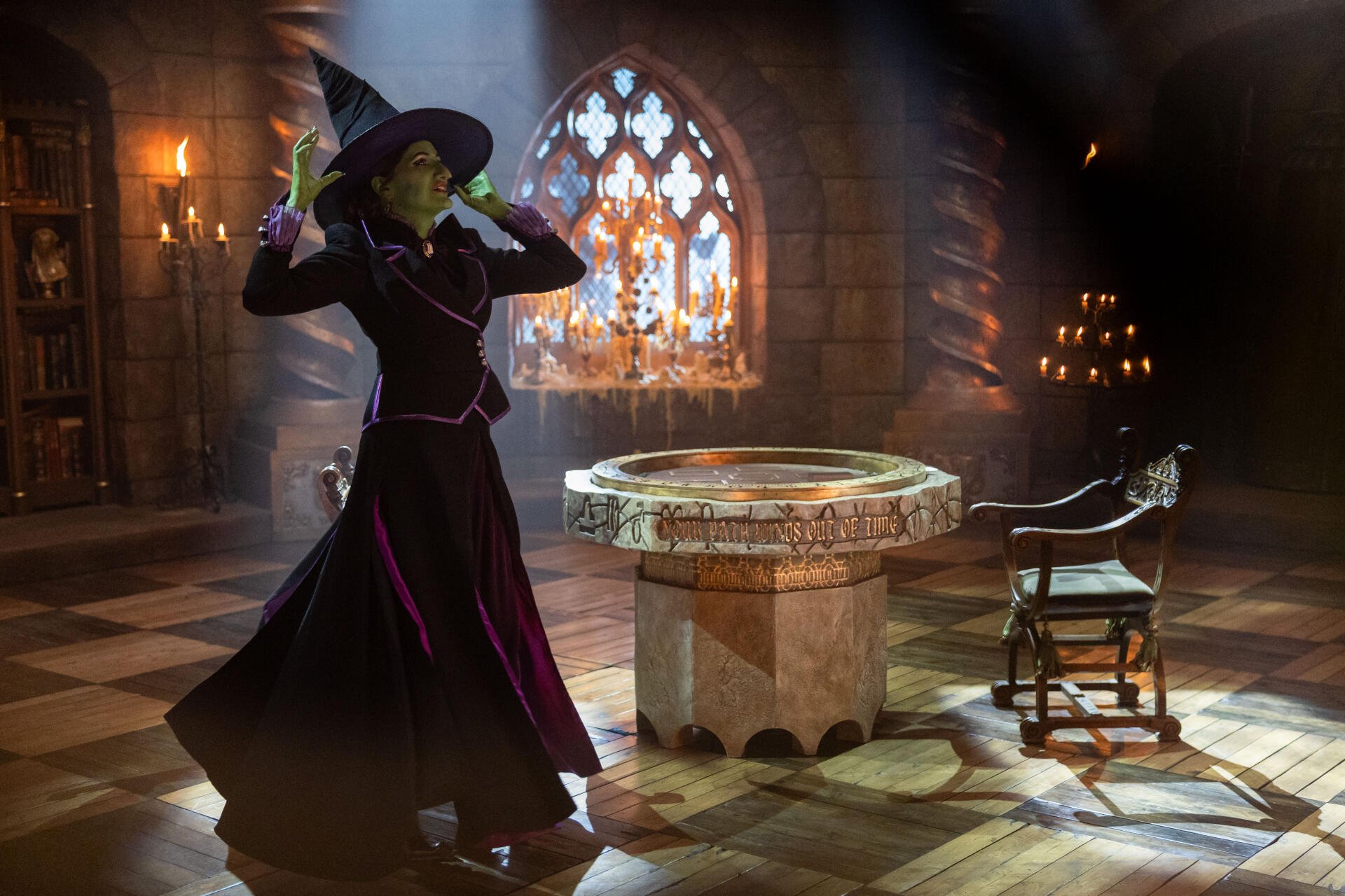A witch in a black and purple outfit strikes a dramatic pose in a dimly lit room with a stone table, capturing a scene inspired by the TV show Agatha All Along. 4K Ultra HD wallpaper.