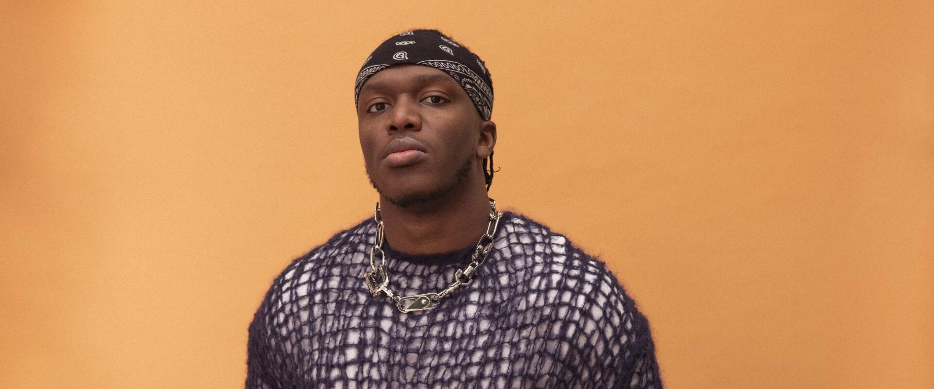 A high-definition desktop wallpaper featuring celebrity KSI, confidently posing against an orange background, wearing a distinctive outfit and accessories.