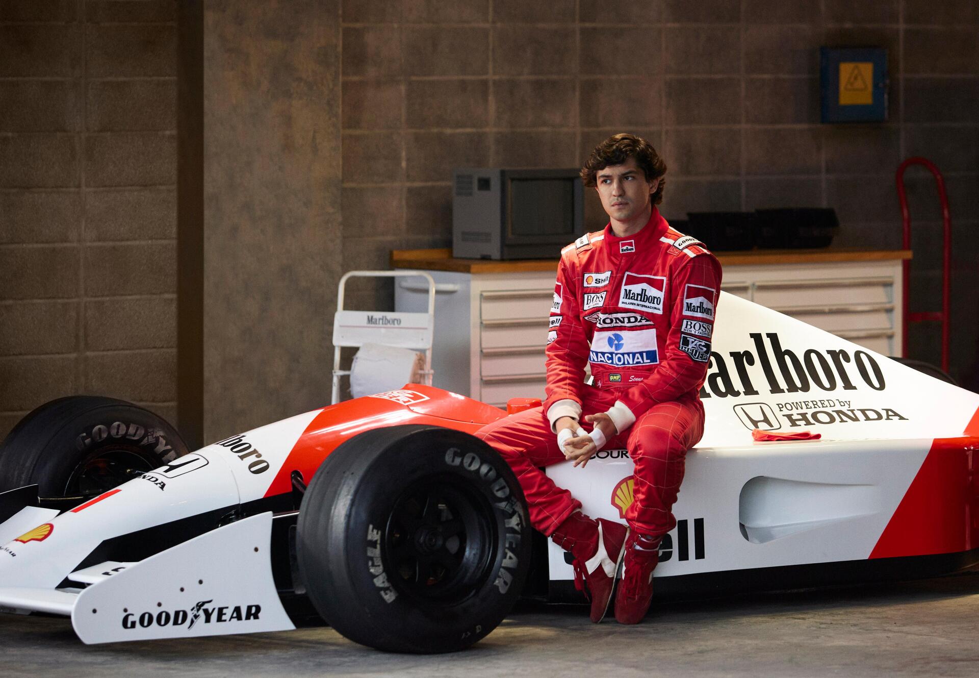 Ayrton Senna sits beside a classic McLaren F1 car in a garage setting. This 4K Ultra HD image captures a moment from the upcoming TV show, Senna (2024).