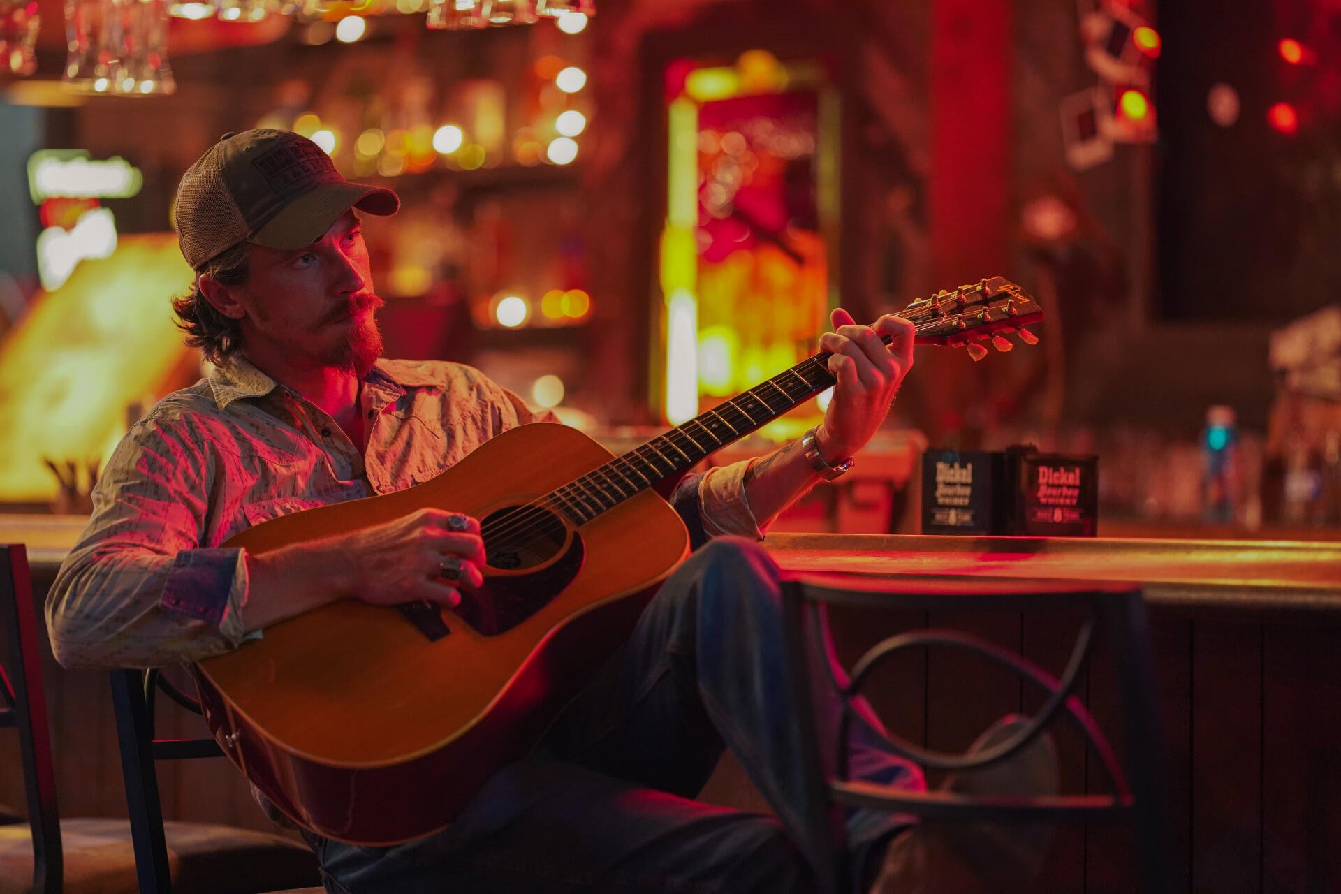 A scene from the TV show Tulsa King, featuring Garrett Hedlund playing acoustic guitar in a vibrant, dimly lit bar, showcasing a lively atmosphere. 4K Ultra HD desktop wallpaper.