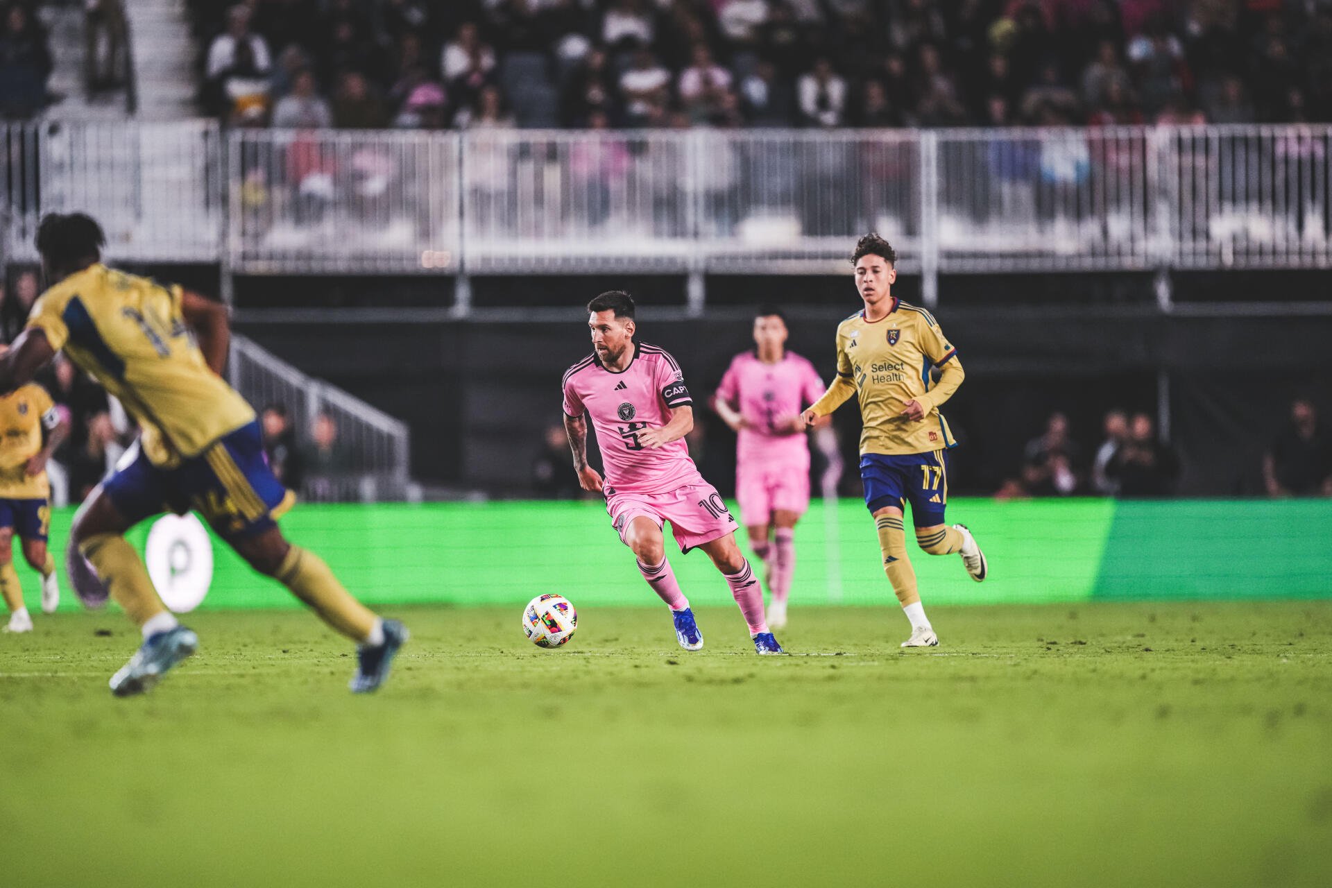 Lionel Messi dribbles the ball in a vibrant pink Inter Miami CF kit during a soccer match, surrounded by opponents on a lively field, captured in stunning 8K Ultra HD.
