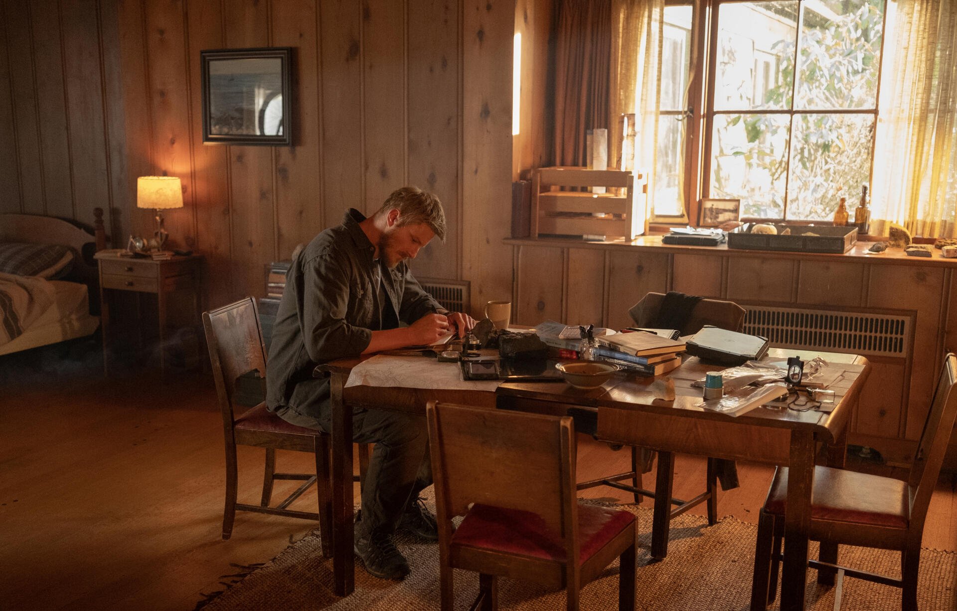 A scene from the TV show Earth Abides, featuring Alexander Ludwig sitting at a table, absorbed in writing amid a cozy, rustic room filled with books and warm sunlight.