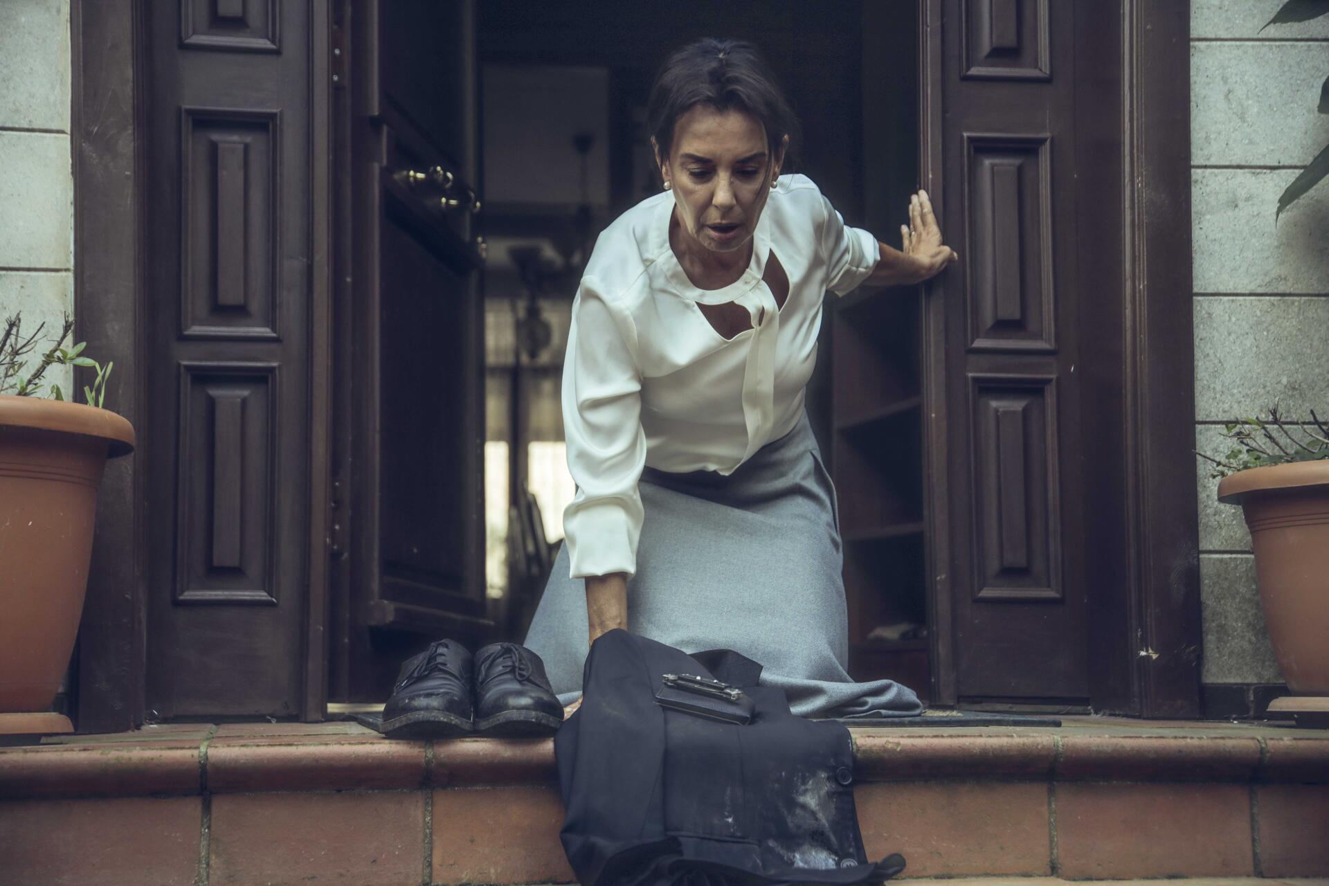 A scene from the TV show The Pit, featuring a woman in a white shirt and gray skirt, struggling as she steps out of a doorway, with a bag and shoes on the steps. 4K Ultra HD wallpaper.