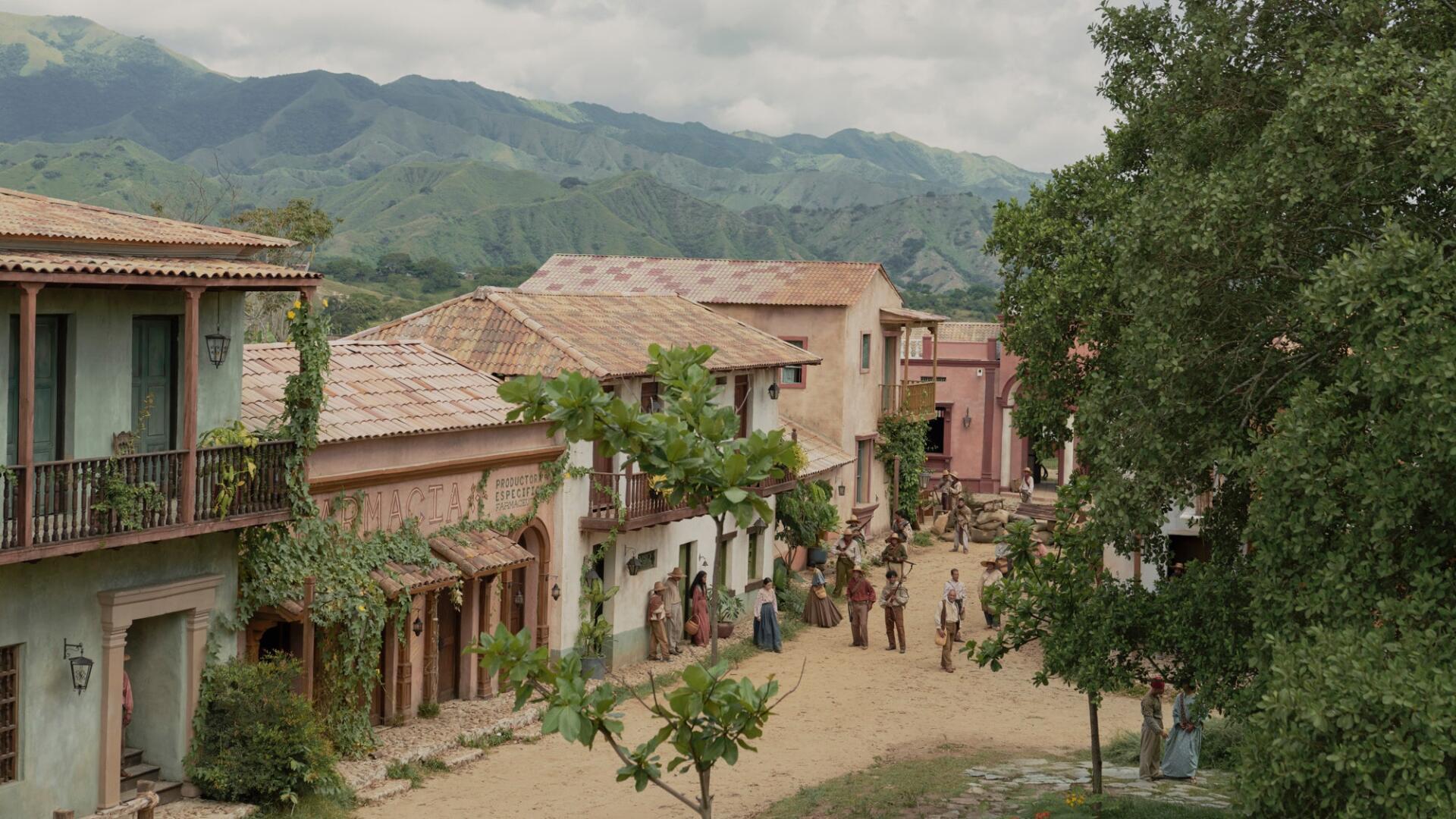 HD PC desktop wallpaper depicting a scenic village scene from the TV show One Hundred Years of Solitude, featuring rustic buildings and lush green mountains.
