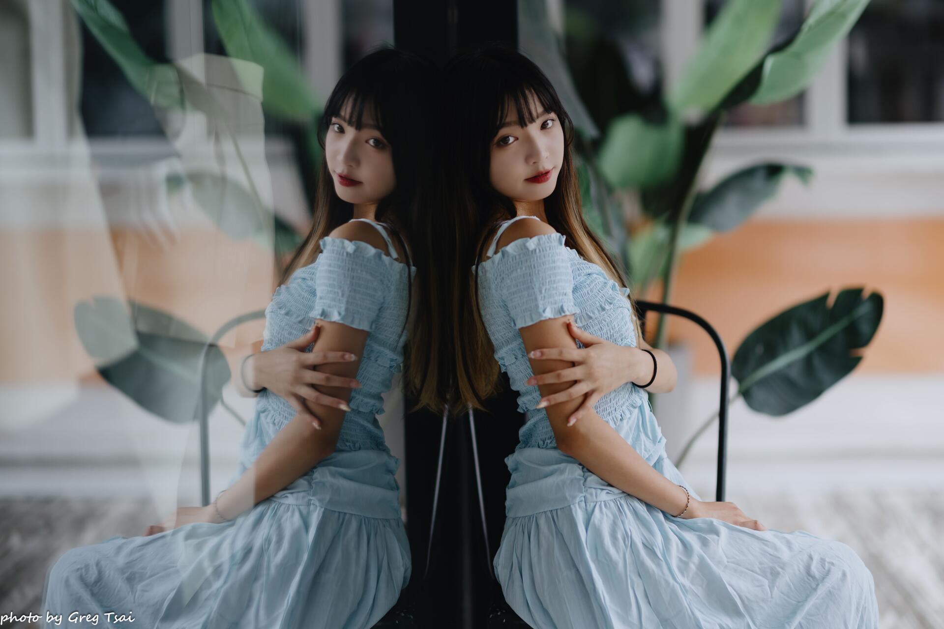 8K Ultra HD PC desktop wallpaper of a woman in a pale blue dress seated by a window, her mirrored reflection on the glass with soft light and large plant leaves in the background.