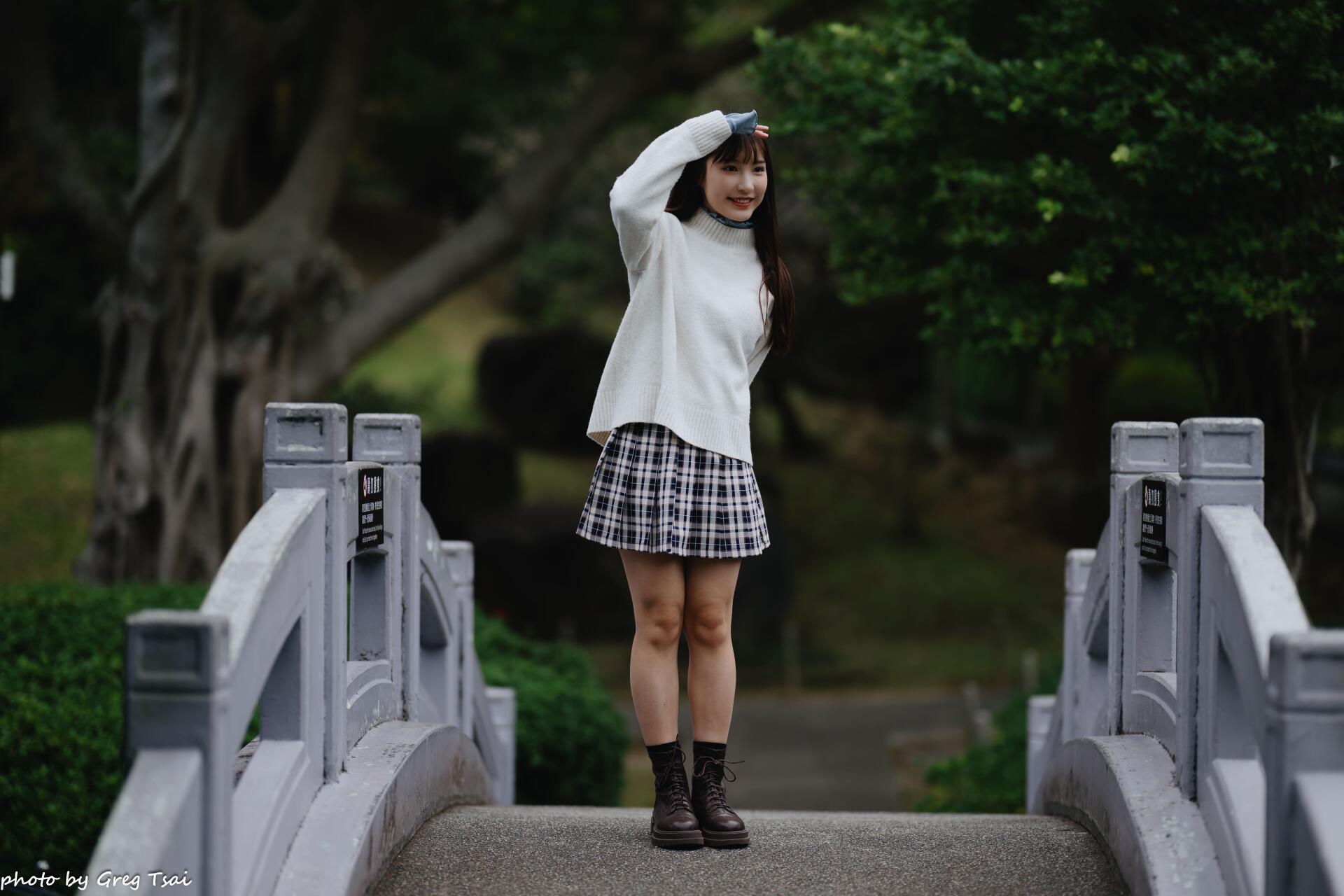 8K Ultra HD PC desktop wallpaper and background: young woman in a white sweater and plaid skirt standing on a small stone bridge, waving amid lush green park trees.