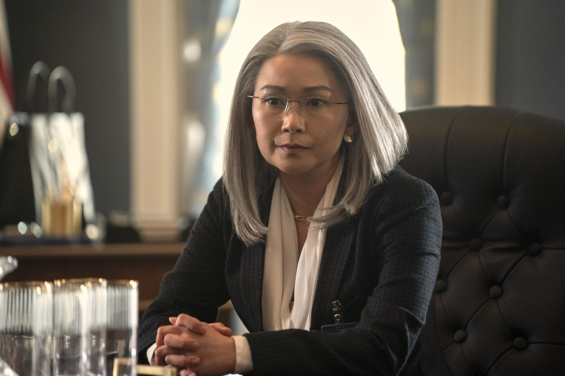 A still from the TV show The Night Agent, featuring Hong Chau with silver hair seated at a desk, showcasing a serious and contemplative expression in a professional setting.