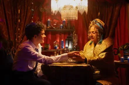 A scene from the TV show Agatha All Along, featuring a young man and an elderly woman in a mystical setting, engaged in a moment of connection amidst rich colors and decor.
