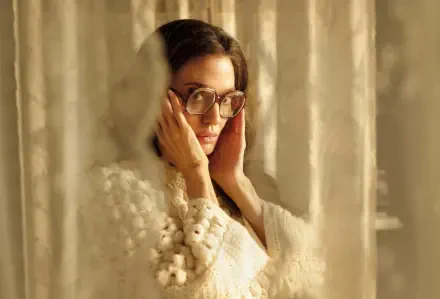 A captivating 4K Ultra HD wallpaper featuring Angelina Jolie as Maria in the 2024 movie, showcasing her thoughtful expression behind delicate curtains.