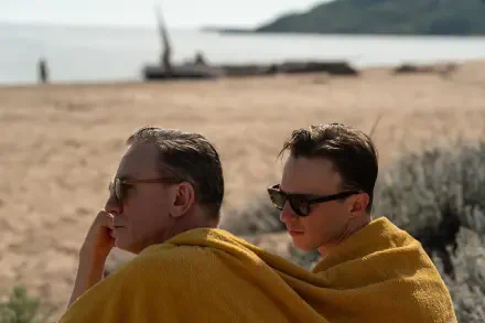 A serene beach scene featuring two men draped in yellow blankets, enjoying time together. The image highlights a moment from a queer-themed film starring Daniel Craig and Drew Starkey.