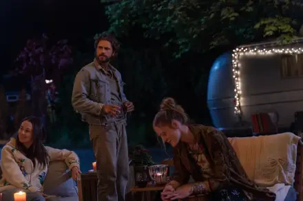 A scene from the TV show *Earth Abides*, featuring three characters gathered around a cozy outdoor setting at night, illuminated by soft lights from a nearby trailer.