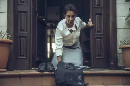 A scene from the TV show The Pit, featuring a woman in a white shirt and gray skirt, struggling as she steps out of a doorway, with a bag and shoes on the steps. 4K Ultra HD wallpaper.