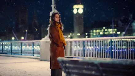 A scene from the TV show Black Doves, featuring Keira Knightley standing by a snowy riverside at night, with a clock tower in the background, creating a serene atmosphere.