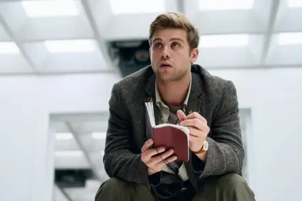 A scene from the TV show Prime Target featuring Leo Woodall, sitting and holding a notebook, with a thoughtful expression in a modern, minimalistic setting. 8K Ultra HD wallpaper.