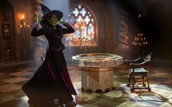 A witch in a black and purple outfit strikes a dramatic pose in a dimly lit room with a stone table, capturing a scene inspired by the TV show Agatha All Along. 4K Ultra HD wallpaper.