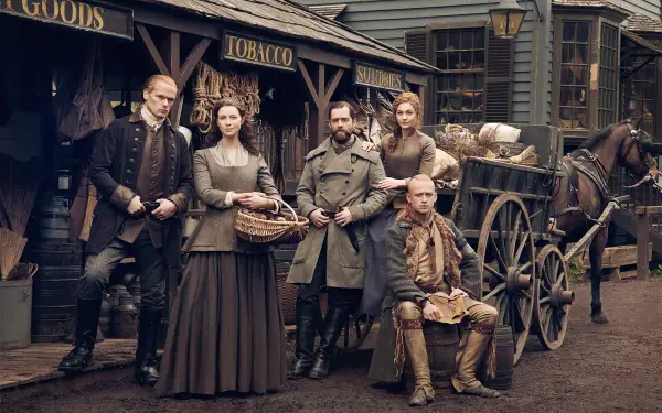 A group of characters from the TV show Outlander poses in a historical setting, showcasing period clothing and a wagon in front of a vintage store.