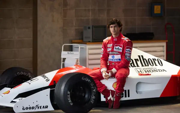 Ayrton Senna sits beside a classic McLaren F1 car in a garage setting. This 4K Ultra HD image captures a moment from the upcoming TV show, Senna (2024).