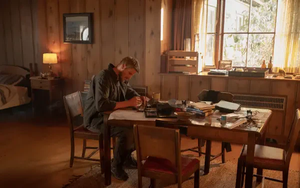 A scene from the TV show Earth Abides, featuring Alexander Ludwig sitting at a table, absorbed in writing amid a cozy, rustic room filled with books and warm sunlight.