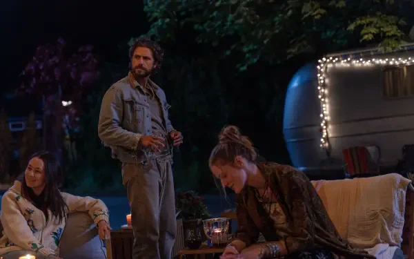A scene from the TV show *Earth Abides*, featuring three characters gathered around a cozy outdoor setting at night, illuminated by soft lights from a nearby trailer.