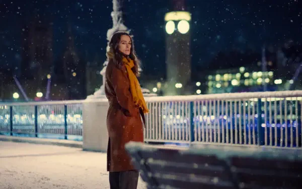 A scene from the TV show Black Doves, featuring Keira Knightley standing by a snowy riverside at night, with a clock tower in the background, creating a serene atmosphere.