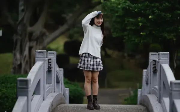 8K Ultra HD PC desktop wallpaper and background: young woman in a white sweater and plaid skirt standing on a small stone bridge, waving amid lush green park trees.