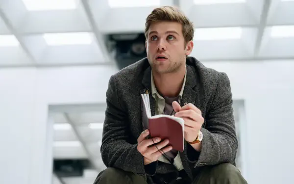 A scene from the TV show Prime Target featuring Leo Woodall, sitting and holding a notebook, with a thoughtful expression in a modern, minimalistic setting. 8K Ultra HD wallpaper.