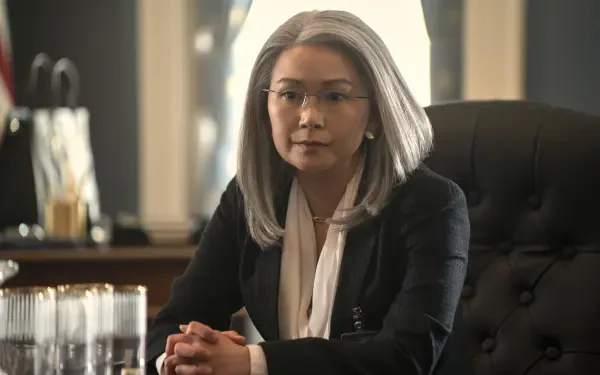 A still from the TV show The Night Agent, featuring Hong Chau with silver hair seated at a desk, showcasing a serious and contemplative expression in a professional setting.