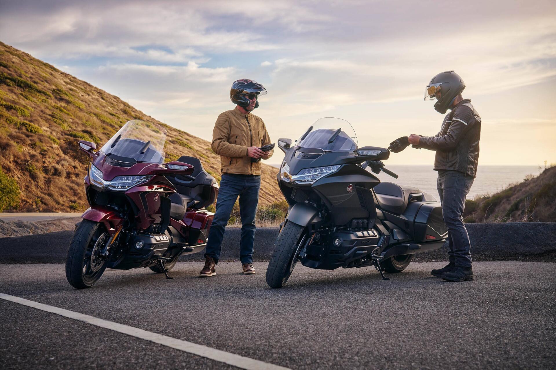 Two riders beside the 25YM Honda GL 1800 Gold Wing motorcycle on a scenic road, with a backdrop of rolling hills and a sunset sky, showcasing an adventure-ready vibe.