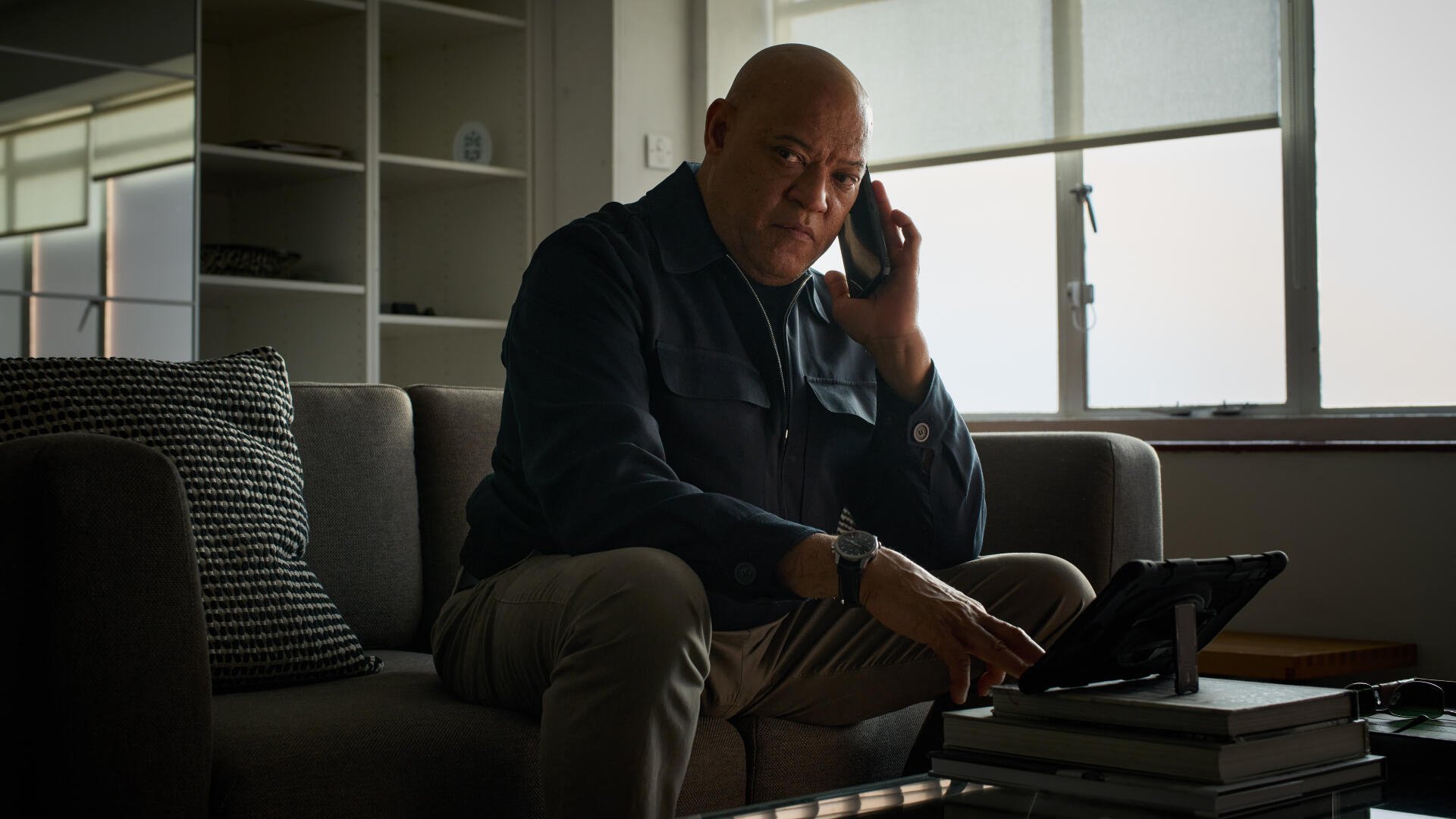 A scene from the movie The Amateur (2025), featuring Laurence Fishburne engaged in a phone conversation, sitting on a sofa with a tablet beside him. 8K Ultra HD quality wallpaper.