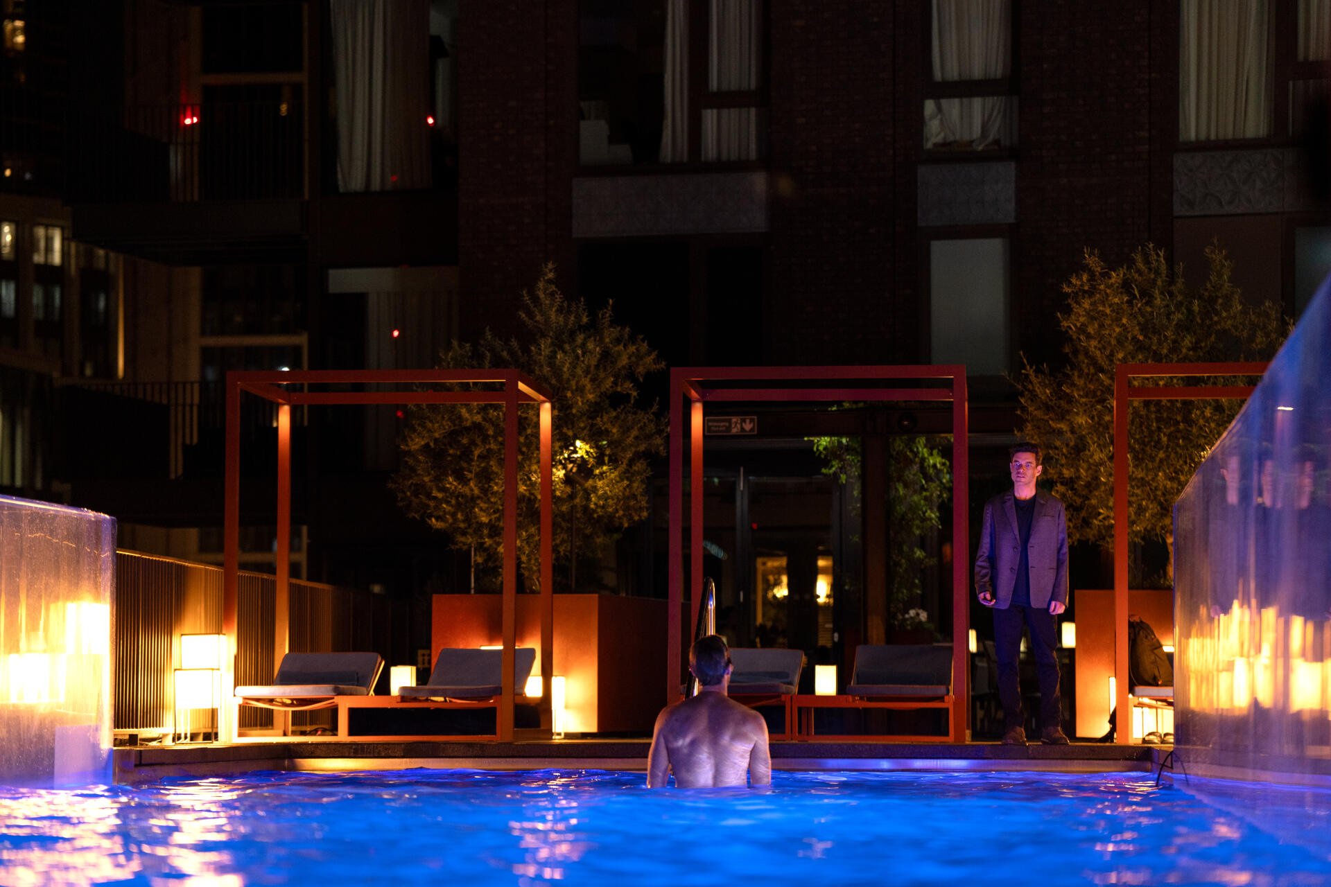 A dramatic scene from the movie The Amateur (2025), featuring a figure in a pool at night, illuminated by ambient lights, with another person standing nearby. 8K Ultra HD resolution.