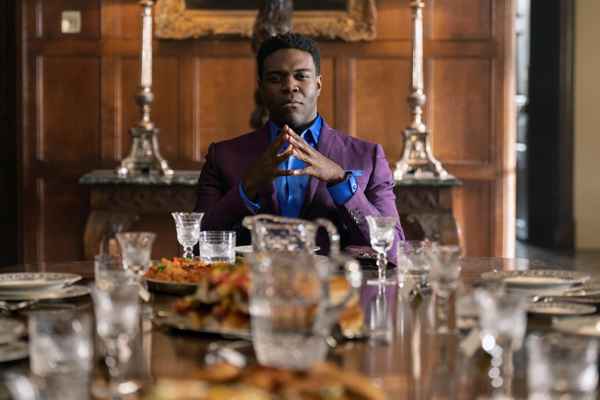 A still from the TV show Ted Lasso, featuring Sam Richardson seated at an elegant dining table, exuding confidence, surrounded by dishes and glasses. 8K Ultra HD quality background.