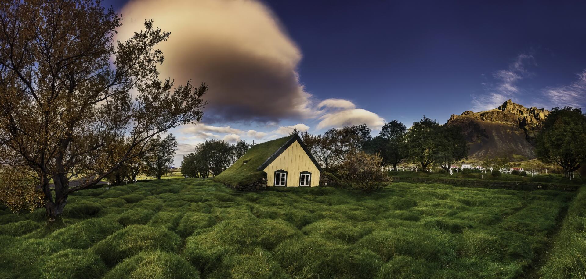 HD desktop wallpaper of an Icelandic landscape featuring a grass-covered cottage near a cemetery and church under a dramatic sky.