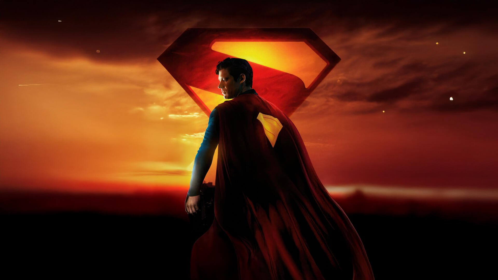 Superman (2025) stands with his cape flowing against a fiery sunset backdrop, with the iconic 'S' symbol glowing behind him in this 4K Ultra HD PC desktop wallpaper.