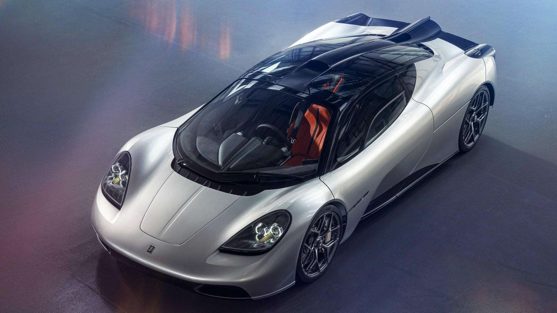 Silver Gordon Murray Automotive hypercar with glass canopy and orange seats, three-quarter top view on an HD PC desktop wallpaper background.