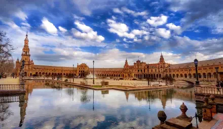 HD PC desktop wallpaper/background: panoramic city scene of ornate historic plaza with arched colonnades and towers reflected in a calm pool under a dramatic blue sky.