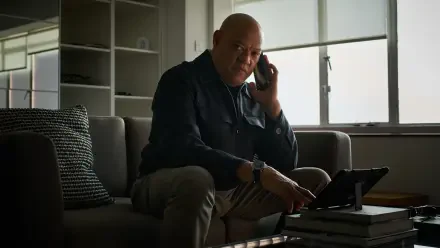 A scene from the movie The Amateur (2025), featuring Laurence Fishburne engaged in a phone conversation, sitting on a sofa with a tablet beside him. 8K Ultra HD quality wallpaper.