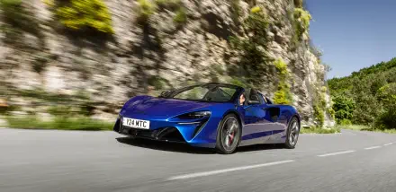 A stunning blue McLaren Artura speeds along a scenic road, showcasing its sleek design against a backdrop of rocky terrain, perfect for a 4K Ultra HD desktop wallpaper.