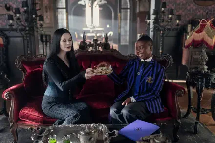 HD PC desktop wallpaper (TV show Wednesday): pale woman in black offers a skull to a smiling student in a blue-striped blazer on a red velvet sofa in a gothic parlor.