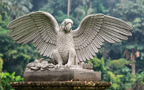 A majestic macaw sculpture with outstretched wings sits atop a pedestal, surrounded by lush greenery, showcased as a stunning 4K Ultra HD desktop wallpaper and background.