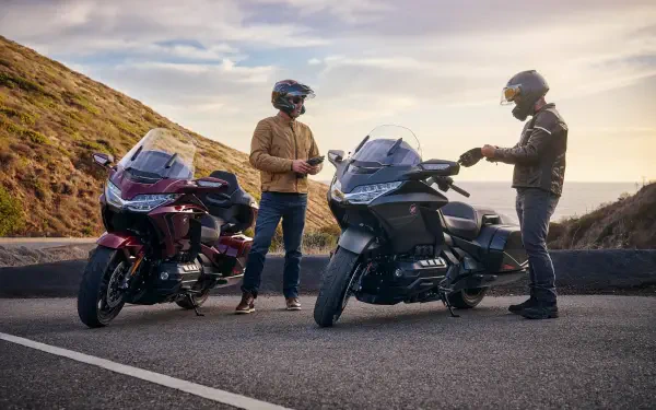 Two riders beside the 25YM Honda GL 1800 Gold Wing motorcycle on a scenic road, with a backdrop of rolling hills and a sunset sky, showcasing an adventure-ready vibe.