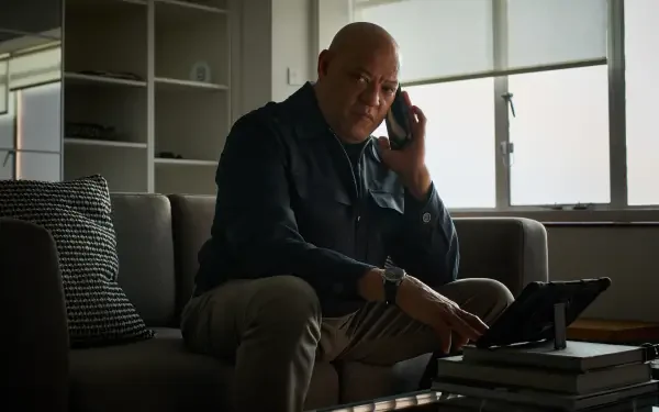 A scene from the movie The Amateur (2025), featuring Laurence Fishburne engaged in a phone conversation, sitting on a sofa with a tablet beside him. 8K Ultra HD quality wallpaper.