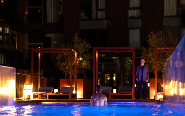 A dramatic scene from the movie The Amateur (2025), featuring a figure in a pool at night, illuminated by ambient lights, with another person standing nearby. 8K Ultra HD resolution.