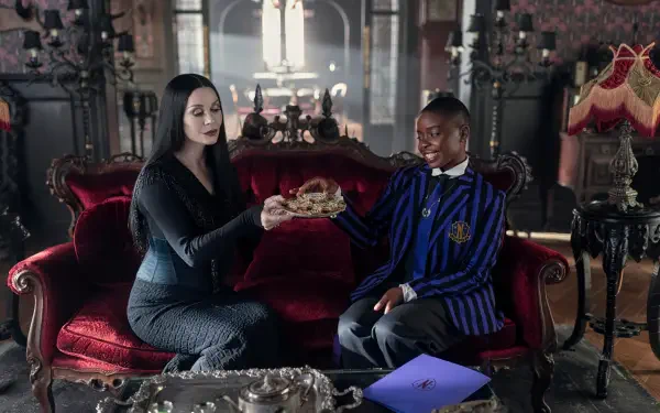 HD PC desktop wallpaper (TV show Wednesday): pale woman in black offers a skull to a smiling student in a blue-striped blazer on a red velvet sofa in a gothic parlor.