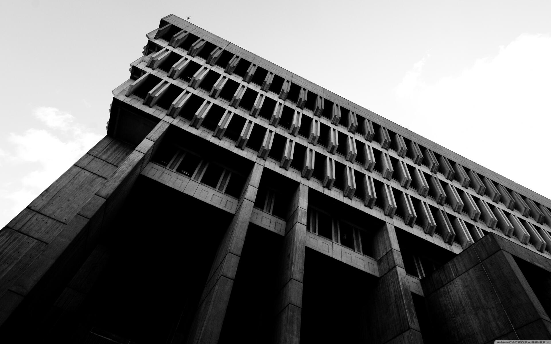 Black-and-white 4K Ultra HD PC desktop wallpaper of a man-made Brutalist architecture building, low-angle view of massive concrete columns and repeating windows against the sky.